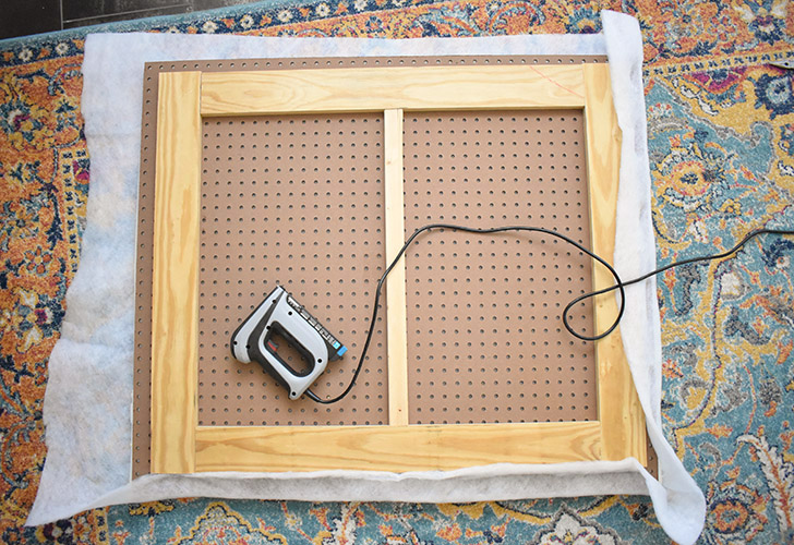 Place quilt batting on your work surface and then the headboard on top with the support wood facing you. Trim so that you have 4-5" of batting on all sides. Fold the batting over and staple in place onto the pegboard. Fold at the corners and use several staples to tack it down with as little bulk as possible. Use sharp scissors to trim away the excess.