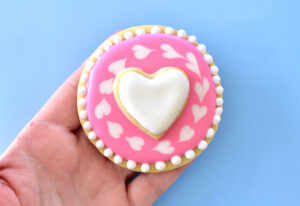For each "O" cookie continue the same process of wet on wet hearts and white beads. Take your small heart shapes and ice in white. Allow both of these cookies plenty of time to dry (overnight is best). Glue the heart into the center of the O shape using a little bit of royal icing onto the back of the heart. Allow an hour or 2 to dry well enough to hold into place before moving.