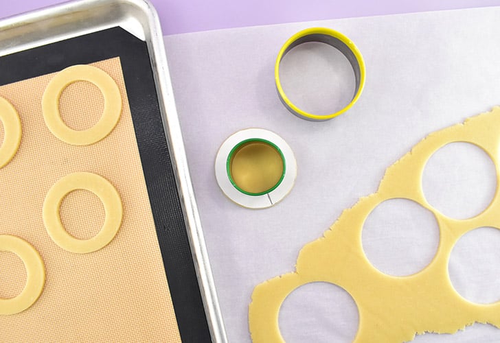 Cut out a 2.6" circle. Gently place your paper guide inside of the cutter and line the mark up with the inside seam of the cutter. Gently pull the cookie cutter straight up, keeping the paper in place as you do. Line up the outer seam of the 1.6" cookie cutter with the line on the guide, making sure the circle fits within the hole and doesn't overlap the paper. Cut the center of the cookie away and place the ring on a silicone mat lined cookie sheet.