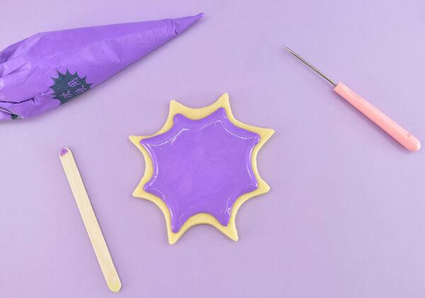 Using your background color of icing pipe following the shape of the cookie making sure it doesn't spill over the sides. Use a toothpick, popsicle stick, or scribe tool to smooth. If the icing is still slightly uneven, gently tap it on your worktop to help level it out.