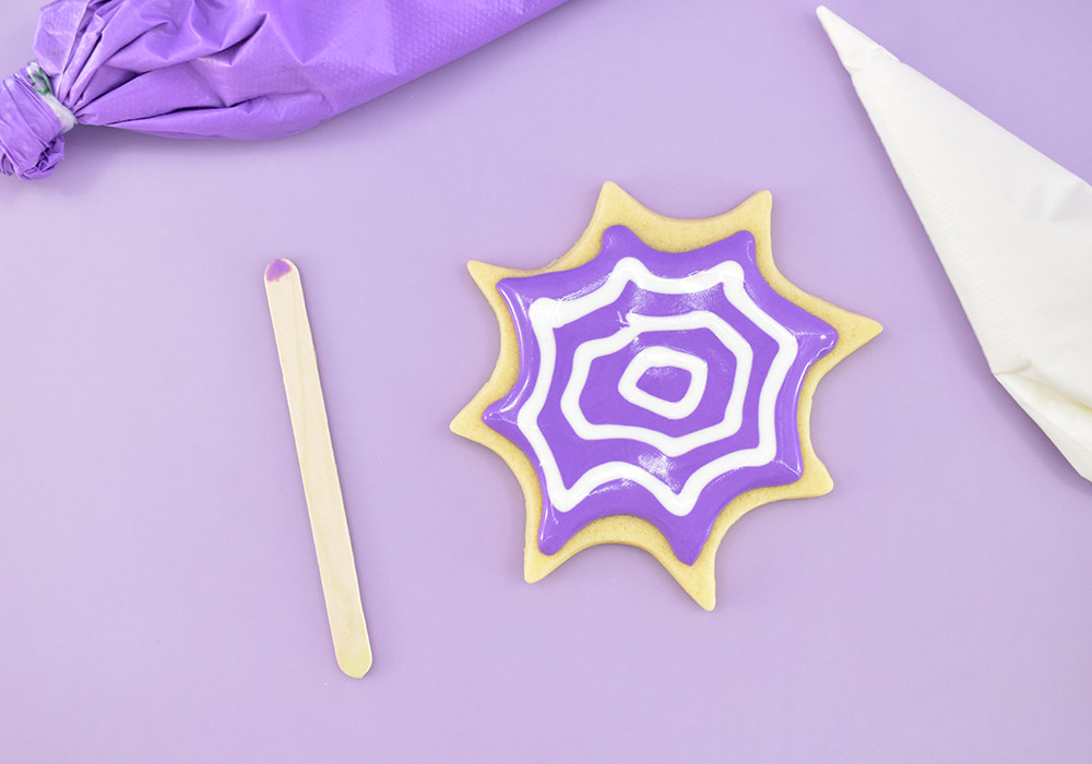 While the background icing is still wet, use your web color to create a series of graduated circles or match the shape of your cookie. Work quickly, you need your icing to remain wet and without crust forming in the icing for the next step.